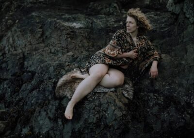 Billie sits on a dark rock, with a sheepskin. Billie is looking off camera, and is holding a dark floral robe closed