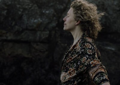 Billie Stands in a dark floral robe with wind blowing Billie's curly blonde hair. The background is dark and rocky.