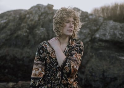 Billie stands and looks off camera, one arm crossed across Billie's chest. Billie is wearing a dark floral robe in front of some rocks.