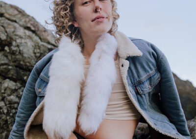 Billie is sitting on some rocks and looking down at the camera. Billie is wearing a denim jacket and fur.